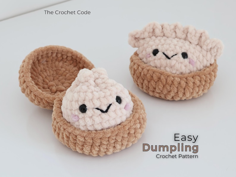 May include: Two crocheted plush dumplings in brown baskets with light pink tops, black embroidered eyes, and small pink cheeks. The text The Crochet Code and Easy Dumpling Crochet Pattern is visible on the white background surrounding the items.