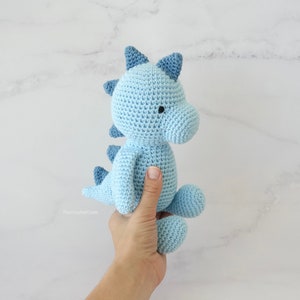 May include: A light blue crocheted dinosaur toy with black eyes. The dinosaur is being held by a hand.