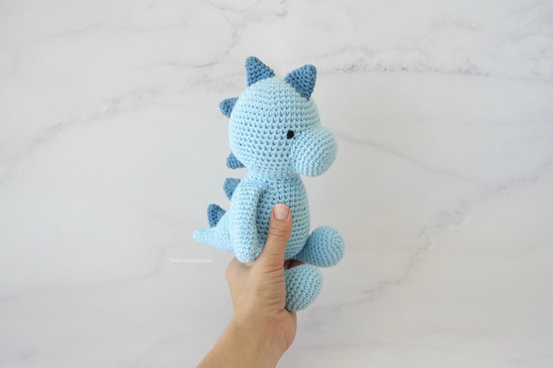 May include: A light blue crocheted dinosaur toy with black eyes. The dinosaur is being held by a hand.