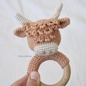 May include: A crocheted brown and white cow rattle with a wooden ring. The rattle is handmade and features a cute cow face with large ears and horns.