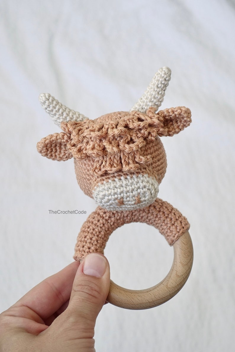 May include: A crocheted brown and white cow rattle with a wooden ring. The rattle is handmade and features a cute cow face with large ears and horns.