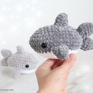 May include: Two gray crocheted shark plushies with white bellies and black eyes. One shark is held in a hand, the other is in the background. The plushies have a soft, textured appearance, and are handmade.