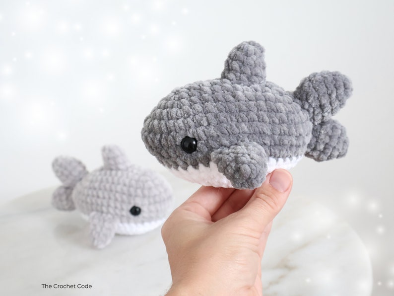 May include: Two gray crocheted shark plushies with white bellies and black eyes. One shark is held in a hand, the other is in the background. The plushies have a soft, textured appearance, and are handmade.