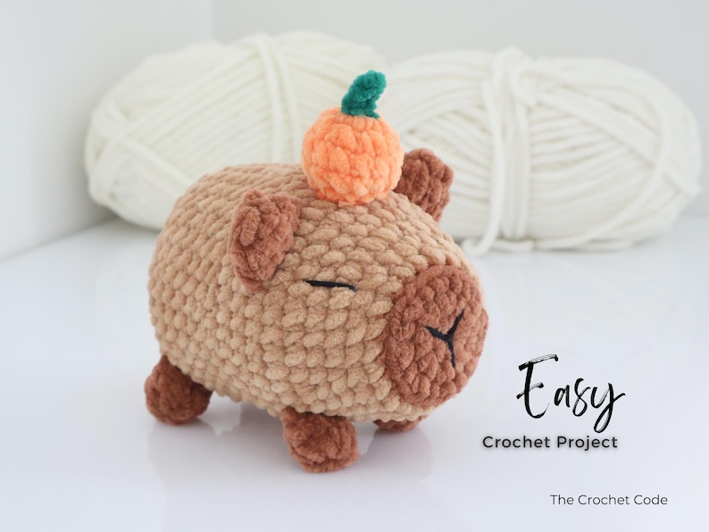 May include: A handmade crochet capybara plushie in shades of brown, with a small orange pumpkin on its head. The plushie has closed eyes and small ears. The text "Easy Crochet Project" is visible.