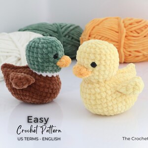 May include: Two crocheted ducklings, one brown and green, the other yellow, sit side-by-side. The ducks have orange beaks and black eyes. The words "Easy Crochet Pattern" are visible. Behind them are balls of yarn in white, green, and orange.