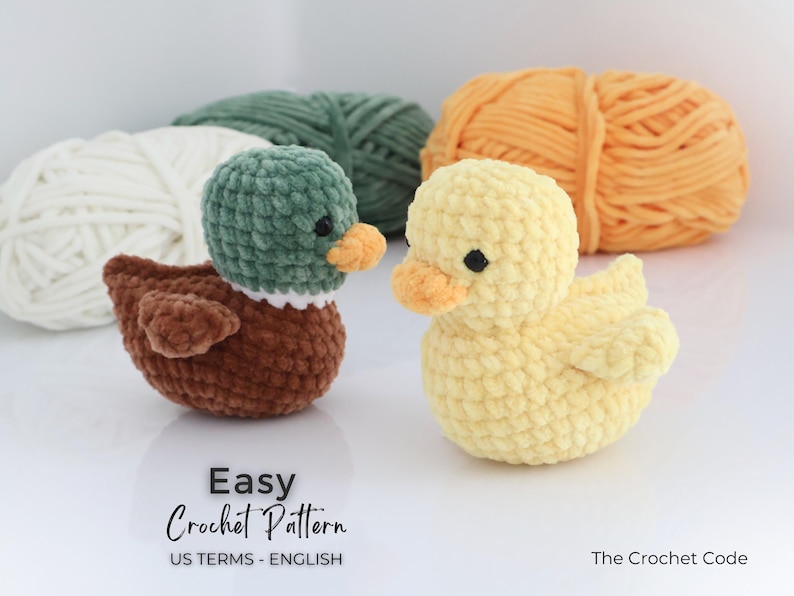 May include: Two crocheted ducklings, one brown and green, the other yellow, sit side-by-side. The ducks have orange beaks and black eyes. The words "Easy Crochet Pattern" are visible. Behind them are balls of yarn in white, green, and orange.