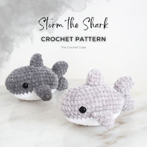 May include: Two crocheted shark toys in shades of gray, with white bellies and black eyes, sit on a marble surface. The text "Storm the Shark" and "CROCHET PATTERN" are displayed above the sharks.