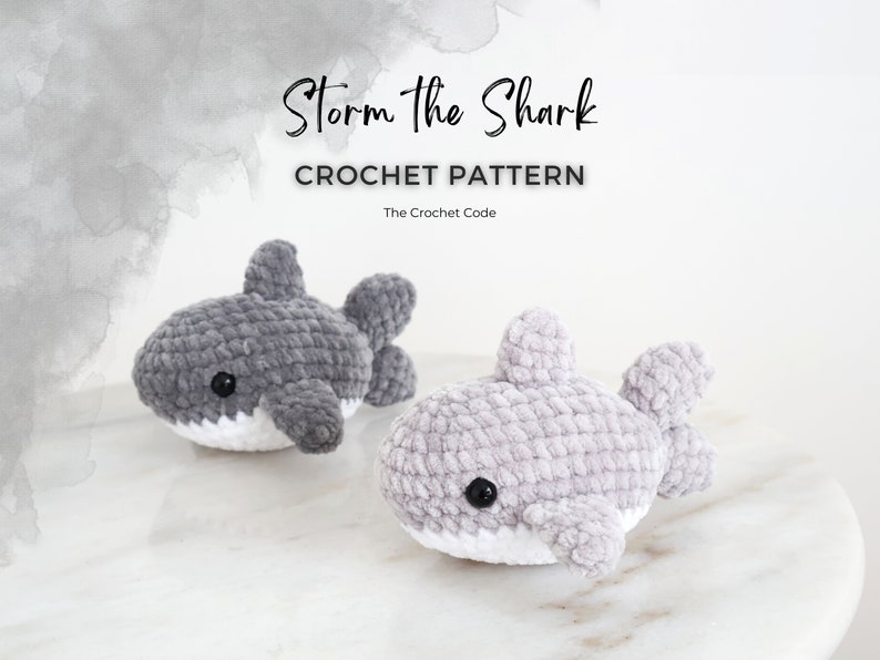 May include: Two crocheted shark toys in shades of gray, with white bellies and black eyes, sit on a marble surface. The text "Storm the Shark" and "CROCHET PATTERN" are displayed above the sharks.