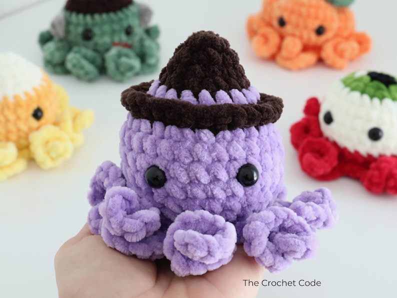 Puede incluir: Un peluche de pulpo tejido a ganchillo de color morado con un sombrero marr&oacute;n oscuro, ojos de pl&aacute;stico negro y tent&aacute;culos rizados. El peluche es parte de una colecci&oacute;n de criaturas tejidas, con el texto The Crochet Code visible abajo.