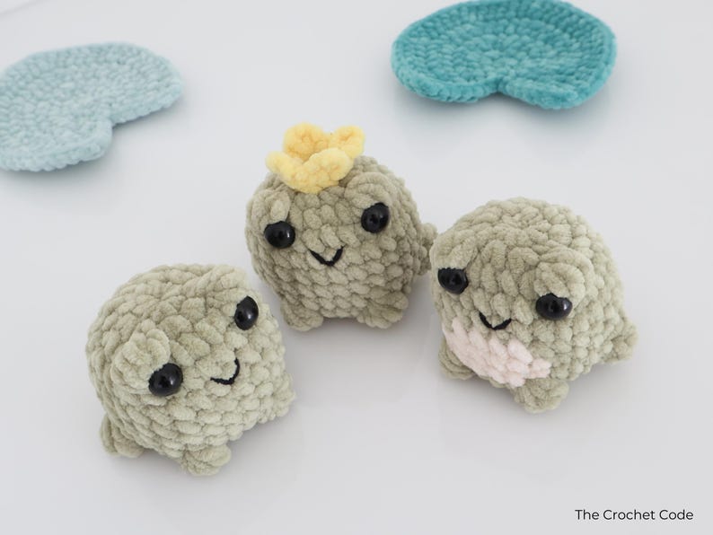 May include: Three handmade crochet creatures with black eyes and smiling faces. One has a yellow flower on its head, and another has a pink patch. Two blue and teal crochet shapes are also in the image. The Crochet Code is in the bottom right corner.