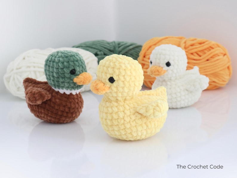 May include: Three crocheted ducklings in brown, yellow, and white, each with an orange beak and black eyes. The ducks are in front of balls of yarn in white, green, and orange. The Crochet Code is written in the bottom right corner.