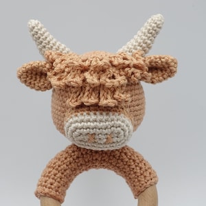 May include: A crocheted brown and white cow rattle toy with a wooden ring. The cow has a fluffy brown mane and white face.