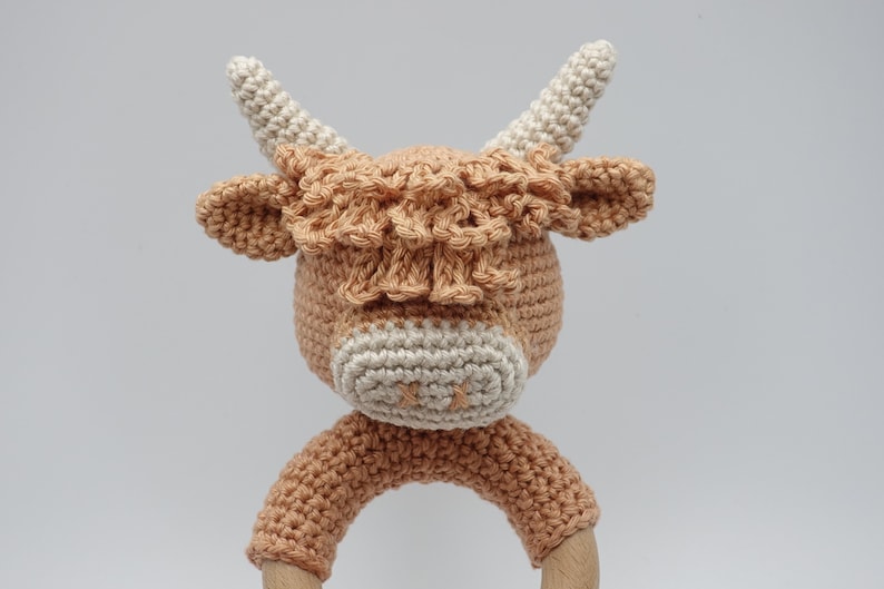 May include: A crocheted brown and white cow rattle toy with a wooden ring. The cow has a fluffy brown mane and white face.