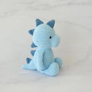 May include: A light blue crocheted dinosaur toy sitting on a white marble surface. The dinosaur has a blue and white spiky back and a small black eye.