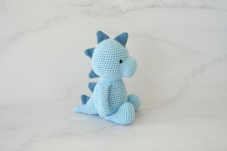 May include: A light blue crocheted dinosaur toy sitting on a white marble surface. The dinosaur has a blue and white spiky back and a small black eye.