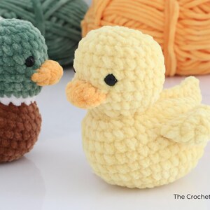 May include: Two handmade crochet ducklings. One is yellow with an orange beak and black eyes. The other duckling is green and brown with a white collar and orange beak. The Crochet Code is written in the bottom right corner.
