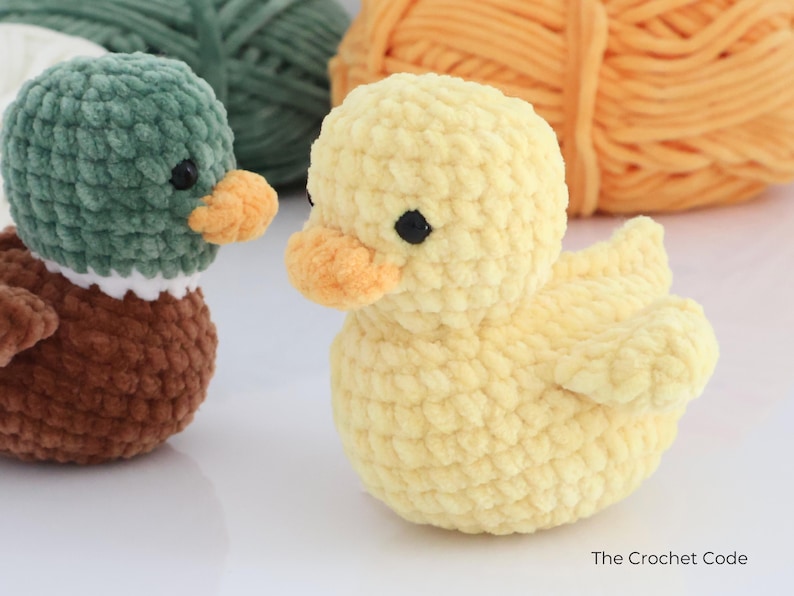 May include: Two handmade crochet ducklings. One is yellow with an orange beak and black eyes. The other duckling is green and brown with a white collar and orange beak. The Crochet Code is written in the bottom right corner.