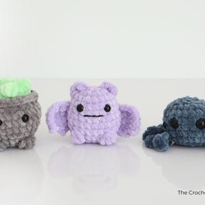May include: Three crocheted plush toys: a grey cauldron with green contents, a lilac bat, and a blue octopus. Each has black button eyes and a stitched mouth. The toys are handmade and sit on a white surface.