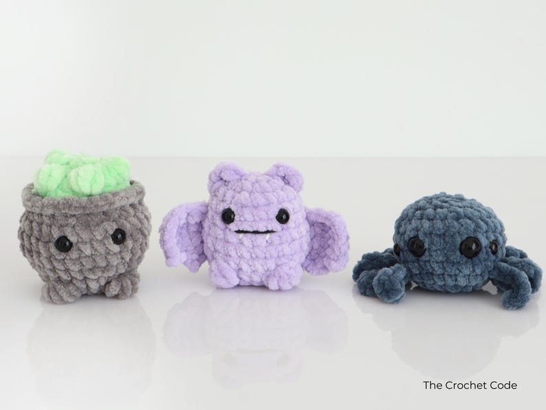 May include: Three crocheted plush toys: a grey cauldron with green contents, a lilac bat, and a blue octopus. Each has black button eyes and a stitched mouth. The toys are handmade and sit on a white surface.