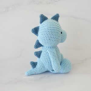 May include: A light blue crocheted dinosaur toy with dark blue accents. The dinosaur is sitting on a white marble surface.