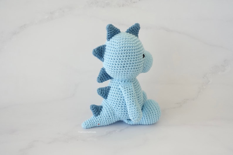 May include: A light blue crocheted dinosaur toy with dark blue accents. The dinosaur is sitting on a white marble surface.