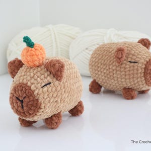 May include: Two handmade crochet capybaras in shades of brown and tan. One has a small orange pumpkin with a green stem on its head. The capybaras have small ears, closed eyes, and are on a white surface.
