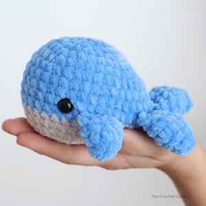 Op de afbeelding: Een handgemaakte, blauwe pluche walvis met een grijze buik en zwarte ogen. De gehaakte walvis is zacht en knuffelig, ideaal voor een kinderspeeltje of kinderkamerdecoratie. De walvis wordt in een hand gehouden.