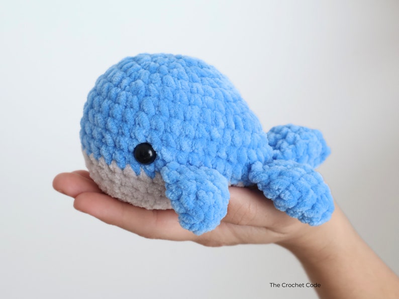 Op de afbeelding: Een handgemaakte, blauwe pluche walvis met een grijze buik en zwarte ogen. De gehaakte walvis is zacht en knuffelig, ideaal voor een kinderspeeltje of kinderkamerdecoratie. De walvis wordt in een hand gehouden.