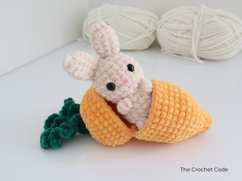 May include: A crocheted bunny toy in a carrot. The bunny is light pink with black eyes and a pink nose. The carrot is orange with green leaves. The Crochet Code is written on the bottom right.