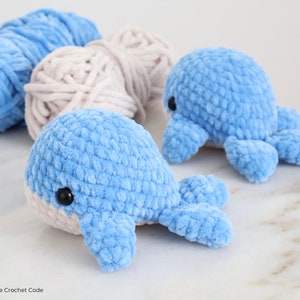 Op de afbeelding: Twee gehaakte speeltjes, een walvis en een octopus, beide lichtblauw met zwarte ogen. Twee bollen garen in lichtblauw en beige op de achtergrond. De speeltjes zijn handgemaakt en hebben een zacht, getextureerd uiterlijk.