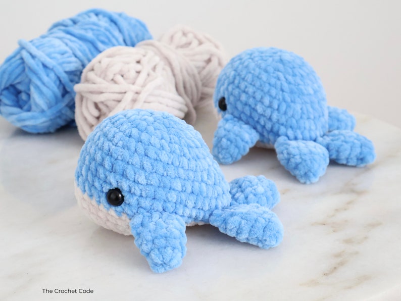 Op de afbeelding: Twee gehaakte speeltjes, een walvis en een octopus, beide lichtblauw met zwarte ogen. Twee bollen garen in lichtblauw en beige op de achtergrond. De speeltjes zijn handgemaakt en hebben een zacht, getextureerd uiterlijk.