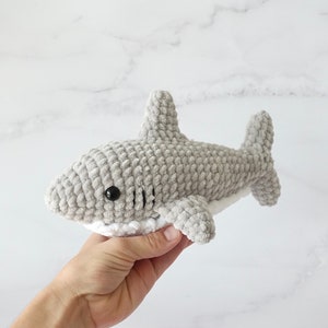 May include: A gray crocheted shark plush toy with white detailing around the mouth and black eyes. The plush toy is being held in a hand.