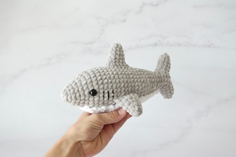 May include: A gray crocheted shark plush toy with white detailing around the mouth and black eyes. The plush toy is being held in a hand.
