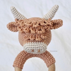 May include: A crocheted brown and white cow rattle toy with a wooden ring. The cow has a long, fluffy mane and a white nose.