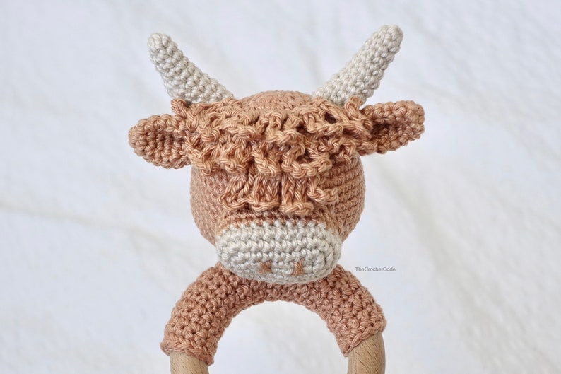 May include: A crocheted brown and white cow rattle toy with a wooden ring. The cow has a long, fluffy mane and a white nose.