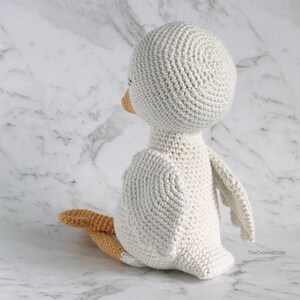 May include: A white crocheted bird with a round head and a small orange beak. The bird is sitting on a white marble surface.