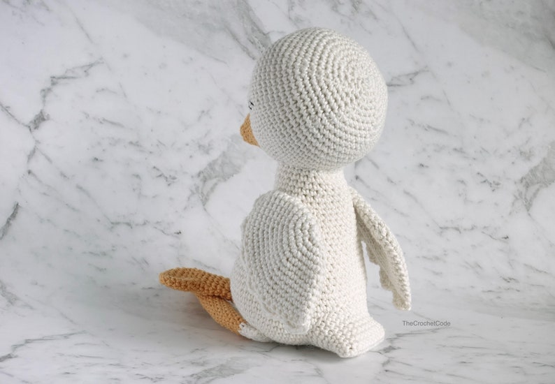 May include: A white crocheted bird with a round head and a small orange beak. The bird is sitting on a white marble surface.