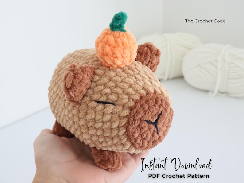 May include: A crocheted capybara plushie in shades of tan and brown, with a small orange pumpkin on its head. The plushie has closed eyes and is shown with the text "The Crochet Code" and "Instant Download PDF Crochet Pattern".