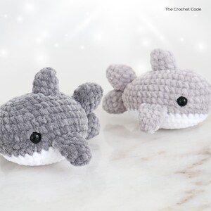 May include: Two gray crochet shark toys with white bellies and black eyes. The sharks have fins and are made of a soft, textured yarn. The Crochet Code is in the top right corner.