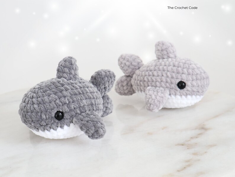 May include: Two gray crochet shark toys with white bellies and black eyes. The sharks have fins and are made of a soft, textured yarn. The Crochet Code is in the top right corner.
