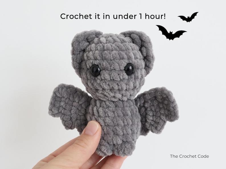 K&ouml;nnte beinhalten: Eine graue geh&auml;kelte Fledermaus mit schwarzen Augen und Fl&uuml;geln wird gehalten. Der Text "Crochet it in under 1 hour!" steht &uuml;ber der Fledermaus. Zwei schwarze Fledermaus-Silhouetten sind im Hintergrund.