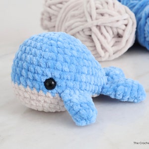 Op de afbeelding: Een handgemaakte blauwe gehaakte walvis met een witte buik en zwart veiligheidsoog. Het pluche speelgoed is gemaakt van zacht, getextureerd garen. Garen in blauwe en grijze tinten staan op de achtergrond.