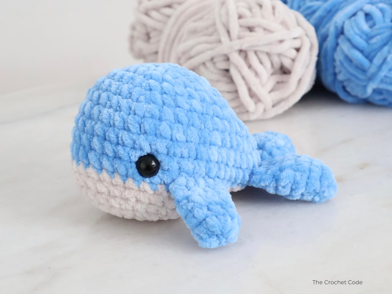Op de afbeelding: Een handgemaakte blauwe gehaakte walvis met een witte buik en zwart veiligheidsoog. Het pluche speelgoed is gemaakt van zacht, getextureerd garen. Garen in blauwe en grijze tinten staan op de achtergrond.