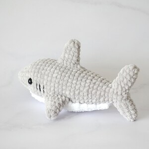 May include: A gray crocheted shark plush toy with white detailing on the underside. The shark has black eyes and a stitched mouth.
