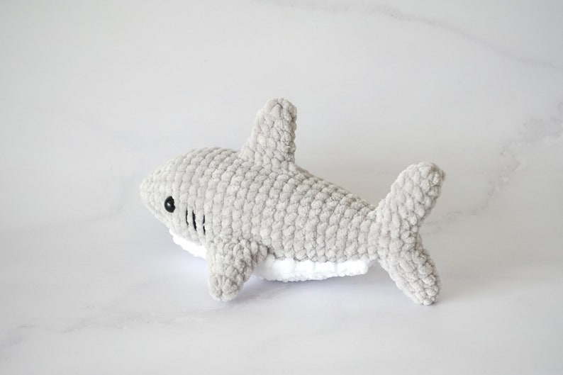 May include: A gray crocheted shark plush toy with white detailing on the underside. The shark has black eyes and a stitched mouth.