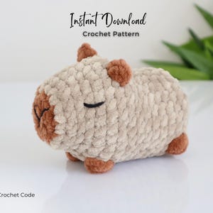 K&ouml;nnte beinhalten: Ein geh&auml;keltes Stofftier, das einem Capybara &auml;hnelt, mit braunen Ohren und Nase. Das Capybara liegt auf der Seite mit geschlossenen Augen. Der Text "Sofortiger Download H&auml;kelanleitung" befindet sich &uuml;ber dem Capybara. Der Text "The Crochet Code" befindet sich unter dem Capybara.