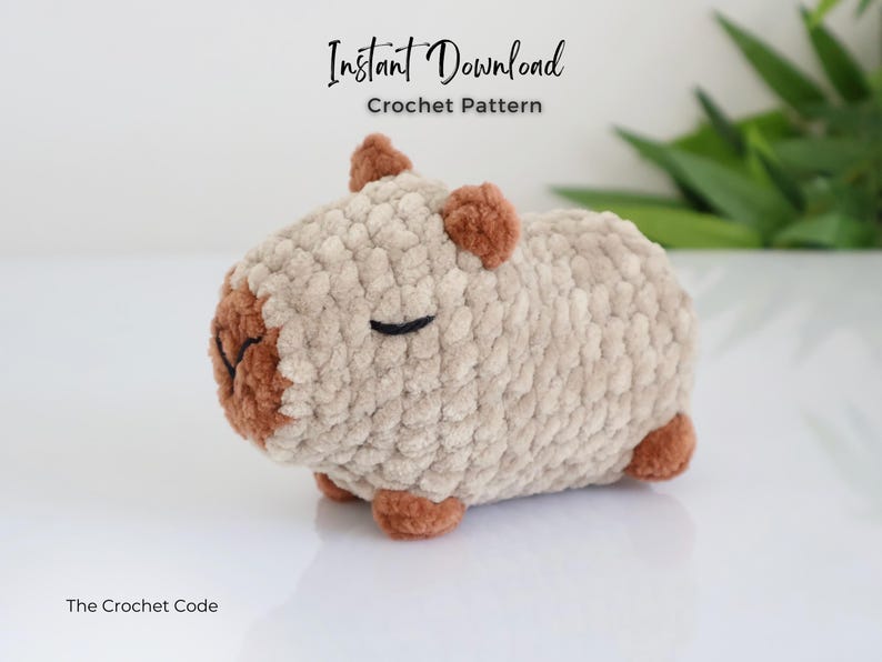 K&ouml;nnte beinhalten: Ein geh&auml;keltes Stofftier, das einem Capybara &auml;hnelt, mit braunen Ohren und Nase. Das Capybara liegt auf der Seite mit geschlossenen Augen. Der Text "Sofortiger Download H&auml;kelanleitung" befindet sich &uuml;ber dem Capybara. Der Text "The Crochet Code" befindet sich unter dem Capybara.