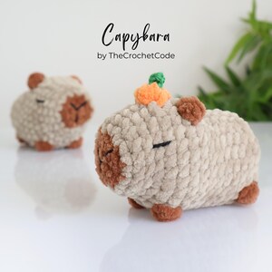 K&ouml;nnte beinhalten: Zwei geh&auml;kelte Capybara-Pl&uuml;schtiere, eines mit einem kleinen orangefarbenen geh&auml;kelten K&uuml;rbis auf dem Kopf. Die Capybaras sind beige mit braunen Akzenten und haben geschlossene Augen. Der Text "Capybara by TheCrochetCode" befindet sich &uuml;ber den Capybaras.