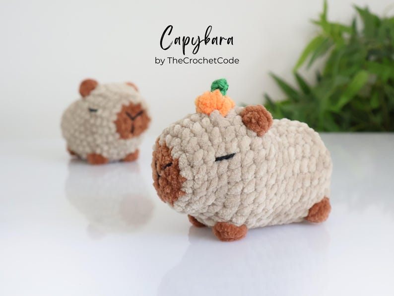 K&ouml;nnte beinhalten: Zwei geh&auml;kelte Capybara-Pl&uuml;schtiere, eines mit einem kleinen orangefarbenen geh&auml;kelten K&uuml;rbis auf dem Kopf. Die Capybaras sind beige mit braunen Akzenten und haben geschlossene Augen. Der Text "Capybara by TheCrochetCode" befindet sich &uuml;ber den Capybaras.