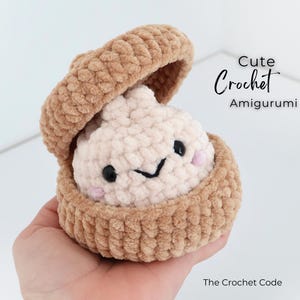 May include: A beige and cream crochet amigurumi dumpling inside a matching beige crochet container. The dumpling has black bead eyes, a stitched black mouth, and pink cheeks. Text reads: Cute Crochet Amigurumi and The Crochet Code.